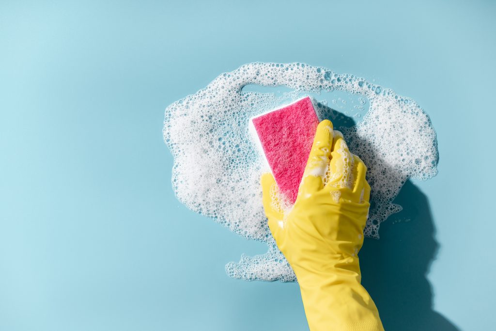 Hand in a yellow rubber glove holds a cleaning sponge and wipes a soapy foam on a blue background. Cleaning concept, cleaning service. Banner. Flat lay, top view